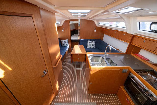 Bright, modern sailboat cabin interior with wood paneling, blue cushioned seating, a small dining table, a galley kitchen with sink and stove, and windows letting in natural light. Berth area visible in the background.