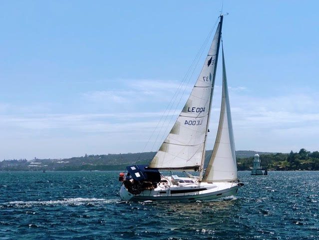 A white sailboat with tan sails glides across a sunny, blue body of water. There is land in the background and a white buoy or small lighthouse on the right. The sky is mostly clear with a few wispy clouds.