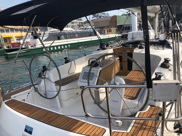 The cockpit of a modern sailboat with twin steering wheels, wooden decking, and digital navigation displays, docked at a marina with a green and white ferry in the background.