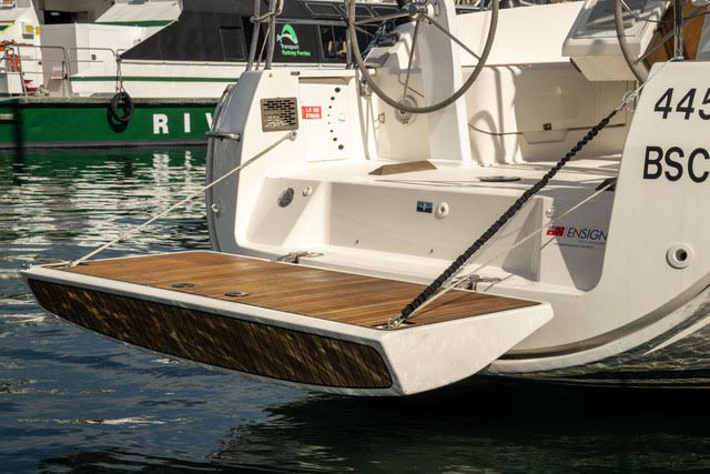 A yacht with its wooden swim platform extended over the water, secured by chains. The stern shows part of the cockpit and helm, with other boats and reflections visible in the background.