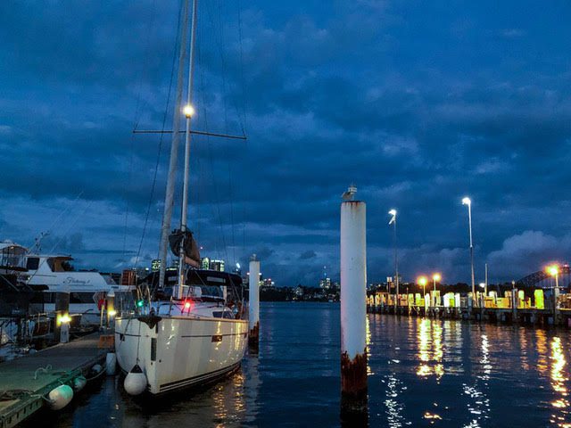 A white sailboat is docked at a marina in the evening, with city lights reflecting on the water and dark clouds in the sky. Bright dock lights illuminate the pier and other boats are visible in the background.