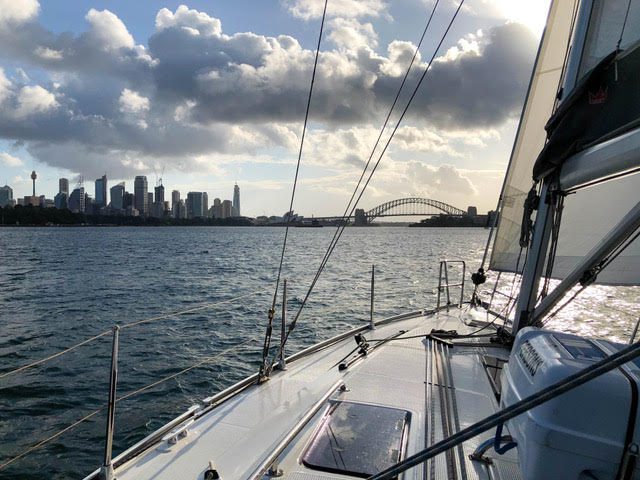 View from a sailboat on the water with the Sydney city skyline and Sydney Harbour Bridge in the distance under a partly cloudy sky, sunlight reflecting off the water and boat deck.
