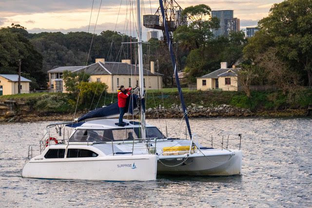 A person in a red jacket stands on the deck of a white catamaran sailboat on a calm body of water, with houses, trees, and city buildings visible in the background.