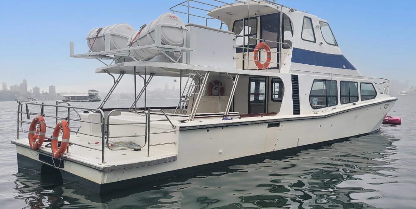 Caitlin’s Yacht, a white boat with blue details, floats on calm water near a hazy city skyline. Two decks, life rings, and rafts are visible—an ideal scene for using your Caitlin's Yacht Ticket.