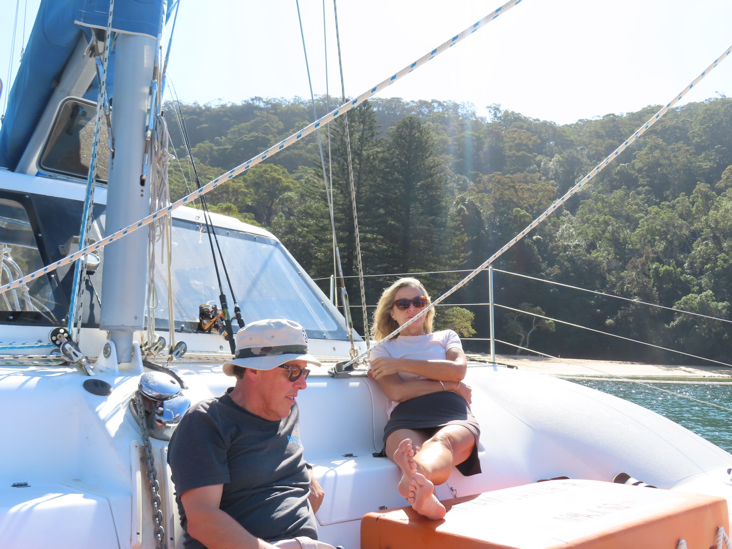 Two people relax on the deck of a sailboat under sunny skies, with forested hills and a sandy beach visible in the background. One person is sitting in a chair while the other reclines with their feet up.