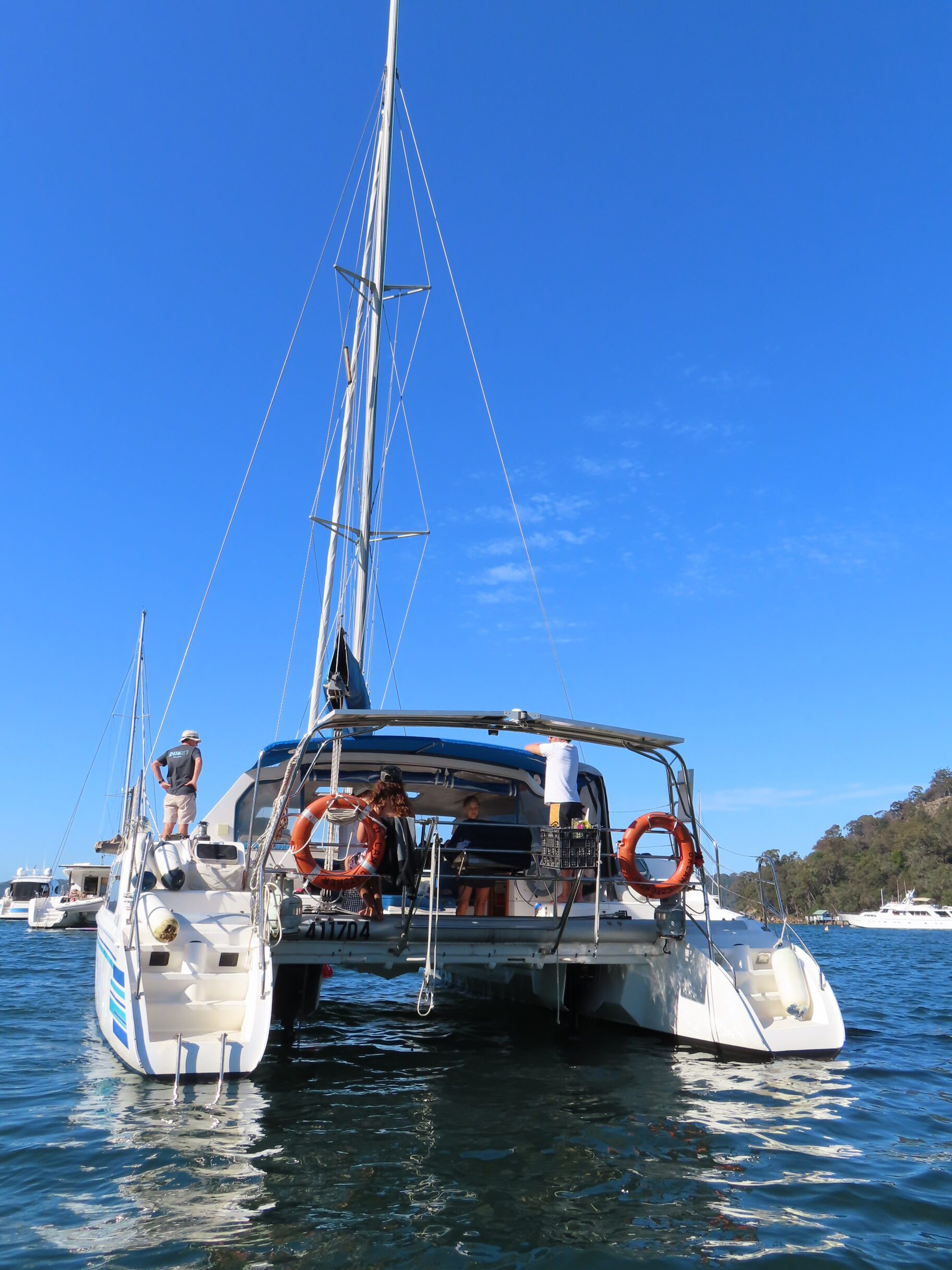 A catamaran with people on board is anchored on calm blue water under a clear sky. Other boats and a tree-lined shore are visible in the background.