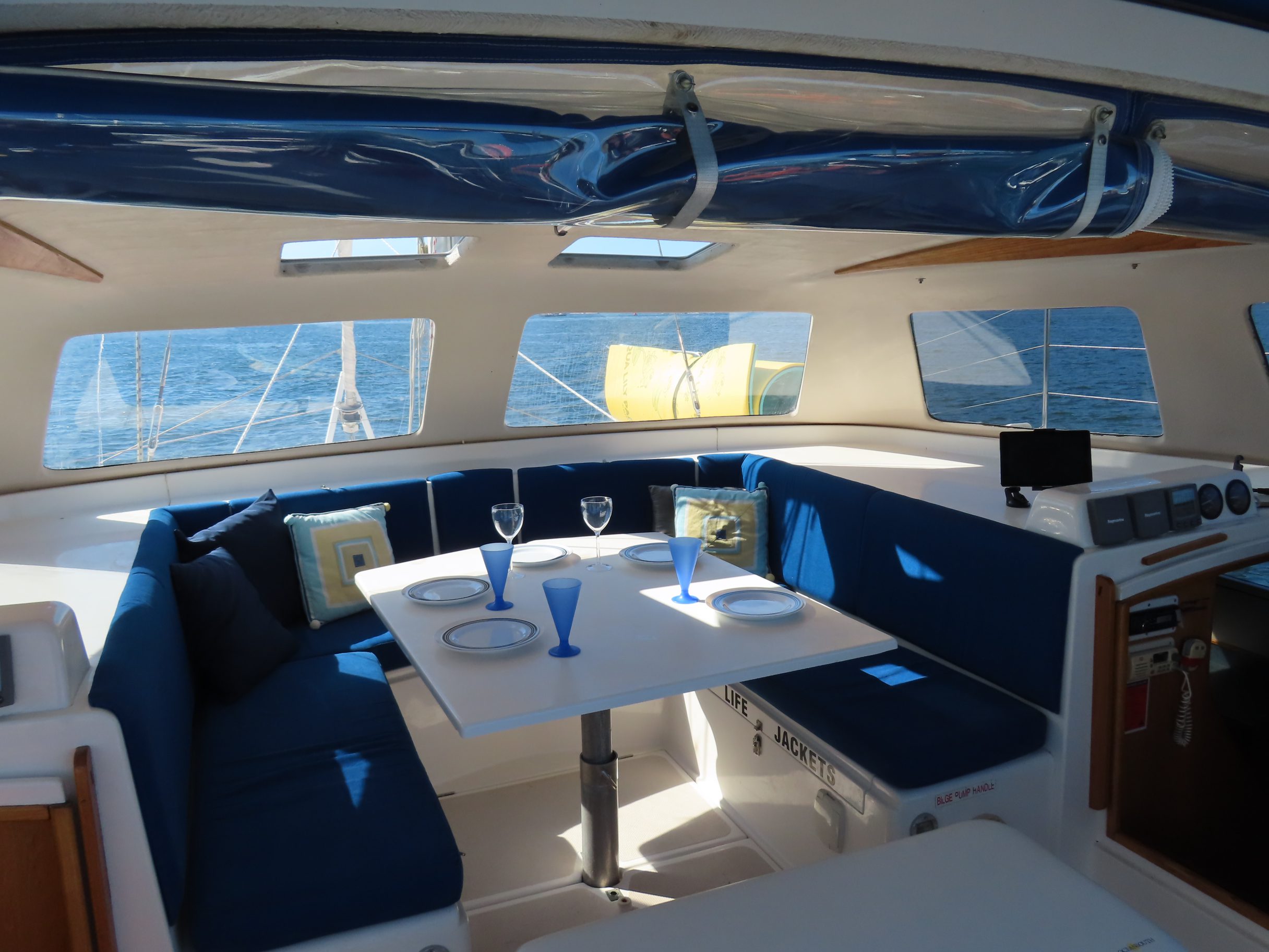 The interior of a sailboat with a dining area featuring blue cushioned seats, a white table set with blue glasses and plates, and large windows showing the ocean outside.