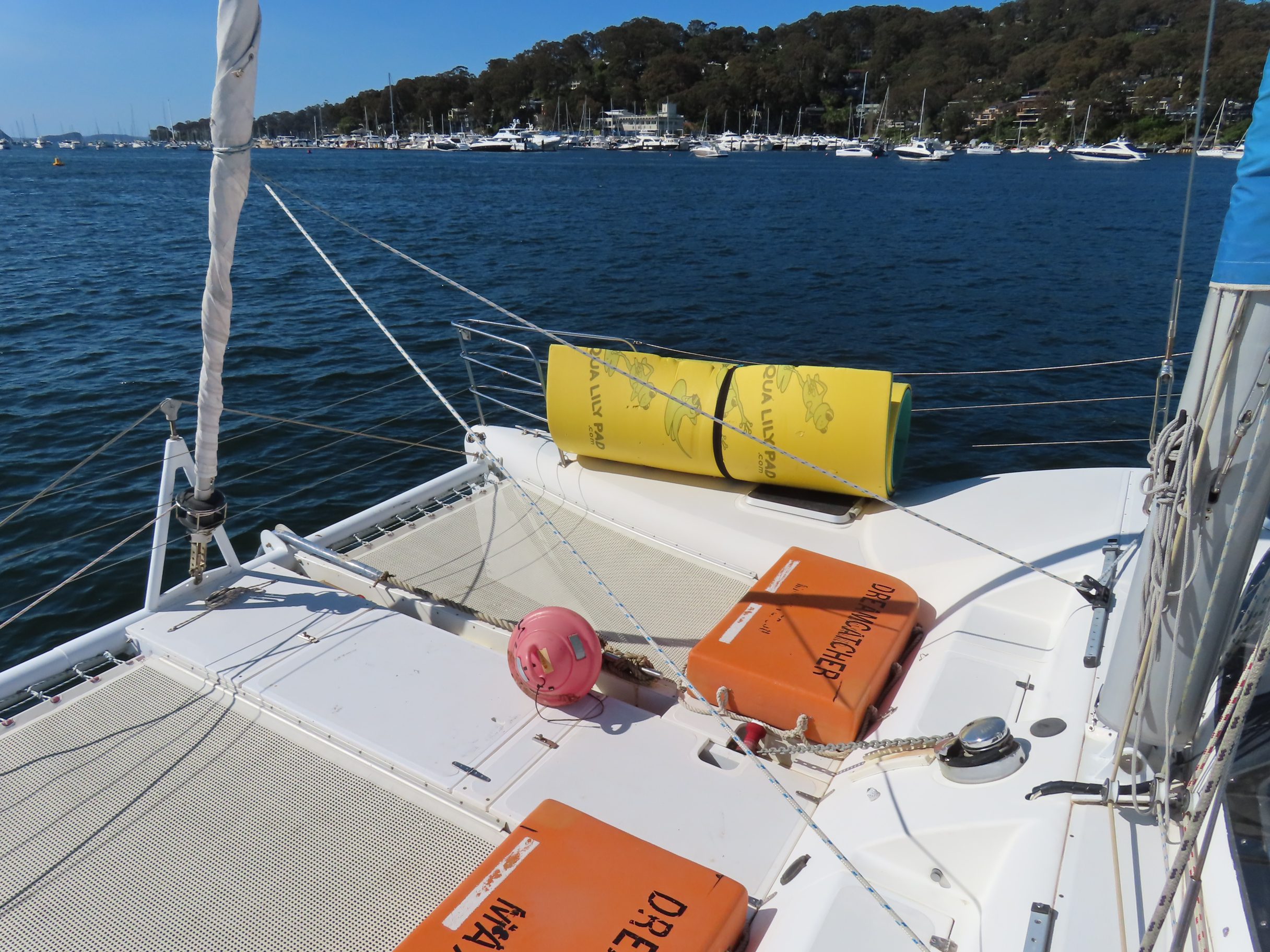 View from the deck of a sailboat with orange life jackets, a pink buoy, and a rolled yellow float. The boat is on blue water near a marina with many docked boats and a tree-covered shoreline in the background.