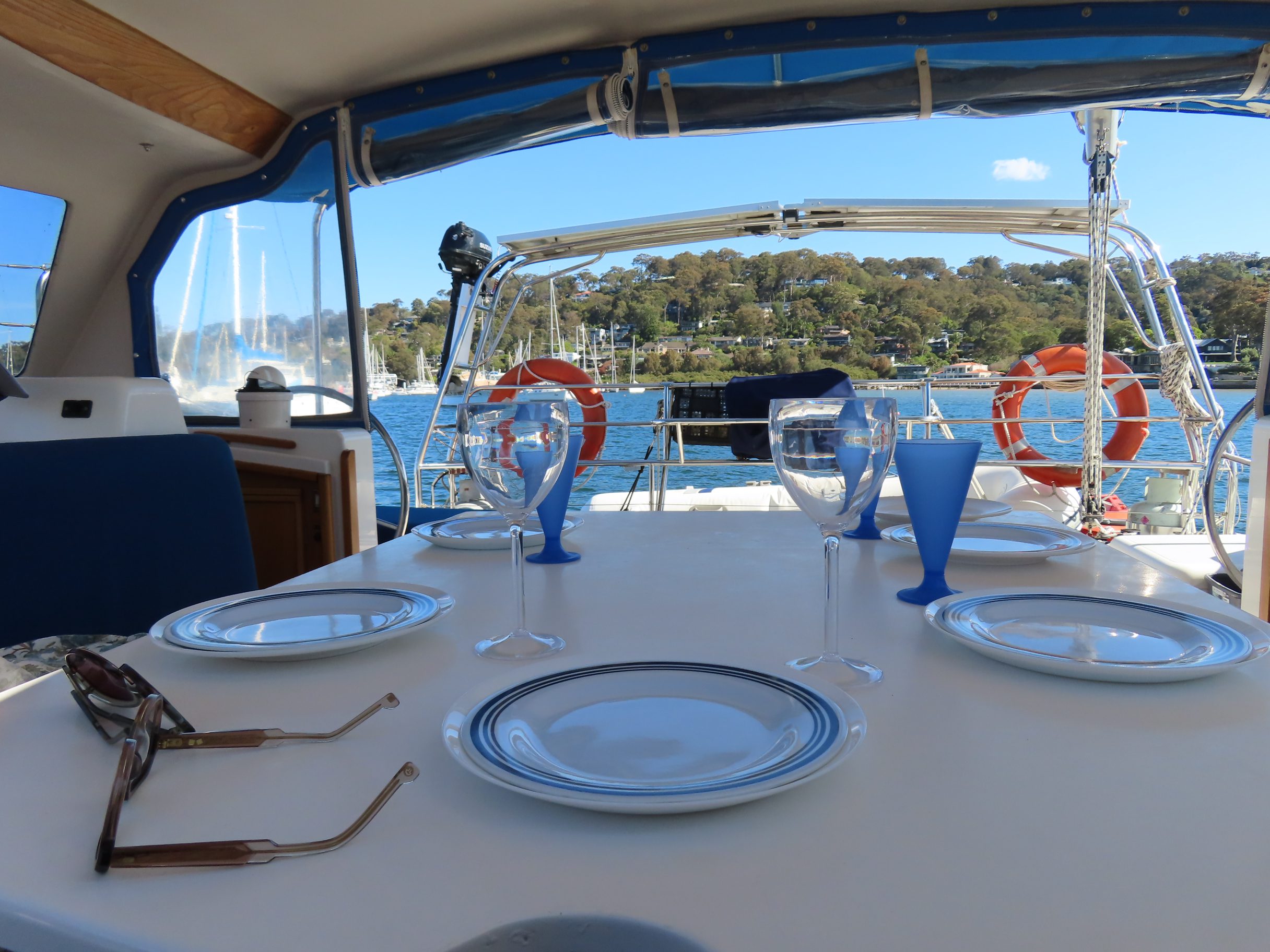 A table set with plates, glasses, and silverware on a boat deck, overlooking calm water and a tree-lined shore in the background. Sunglasses rest on the table near the place settings.