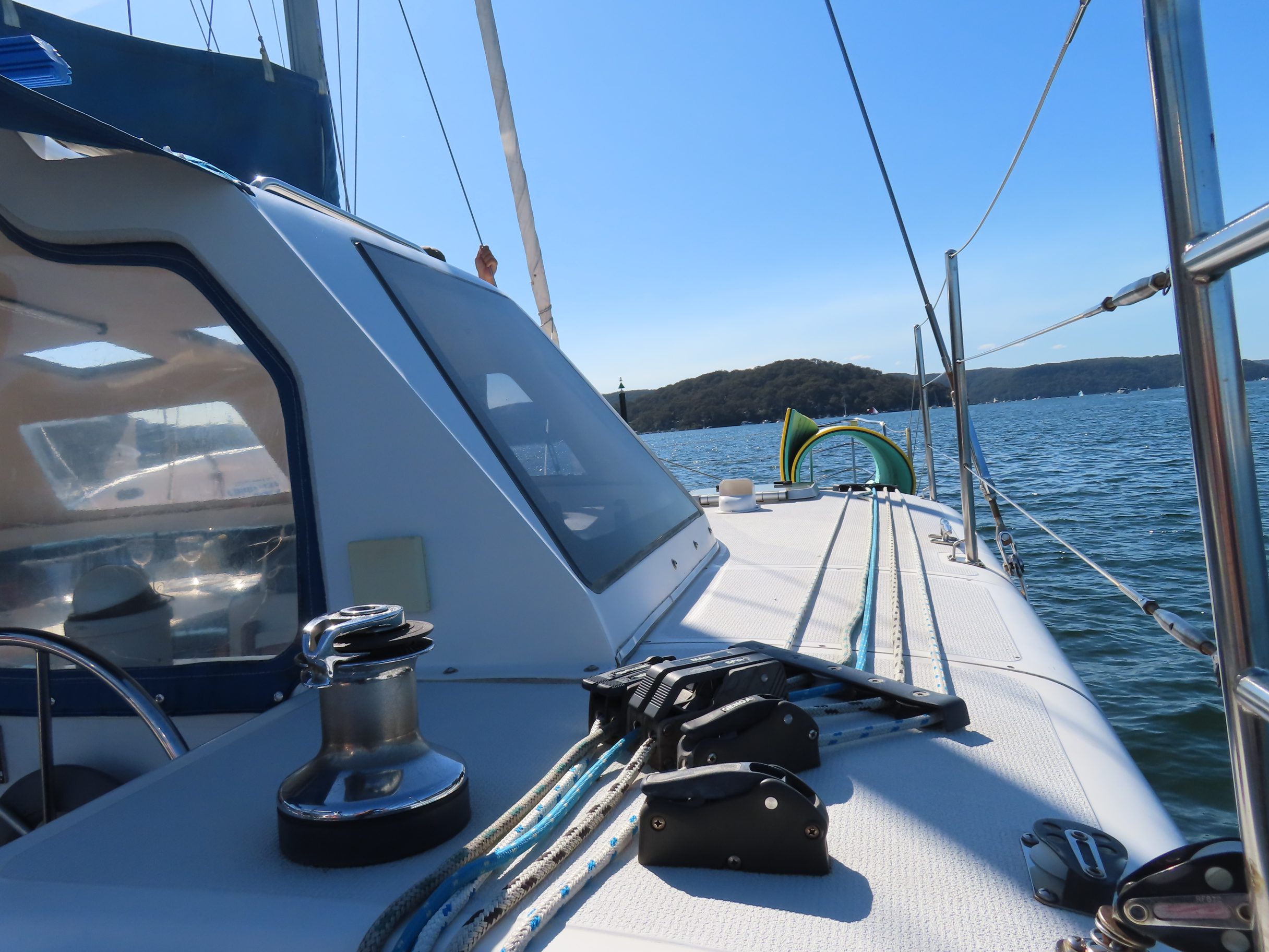 View from the deck of a sailboat on a sunny day, showing ropes, winch, and cabin windows, with blue water and a tree-covered shoreline in the background.