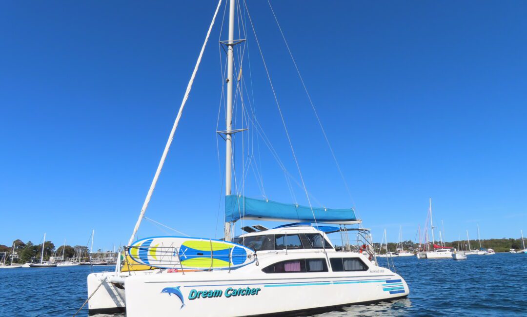 A white catamaran sailboat named "Dream Catcher" floats on calm blue water under a clear sky, with a paddleboard secured on top and other boats visible in the background.