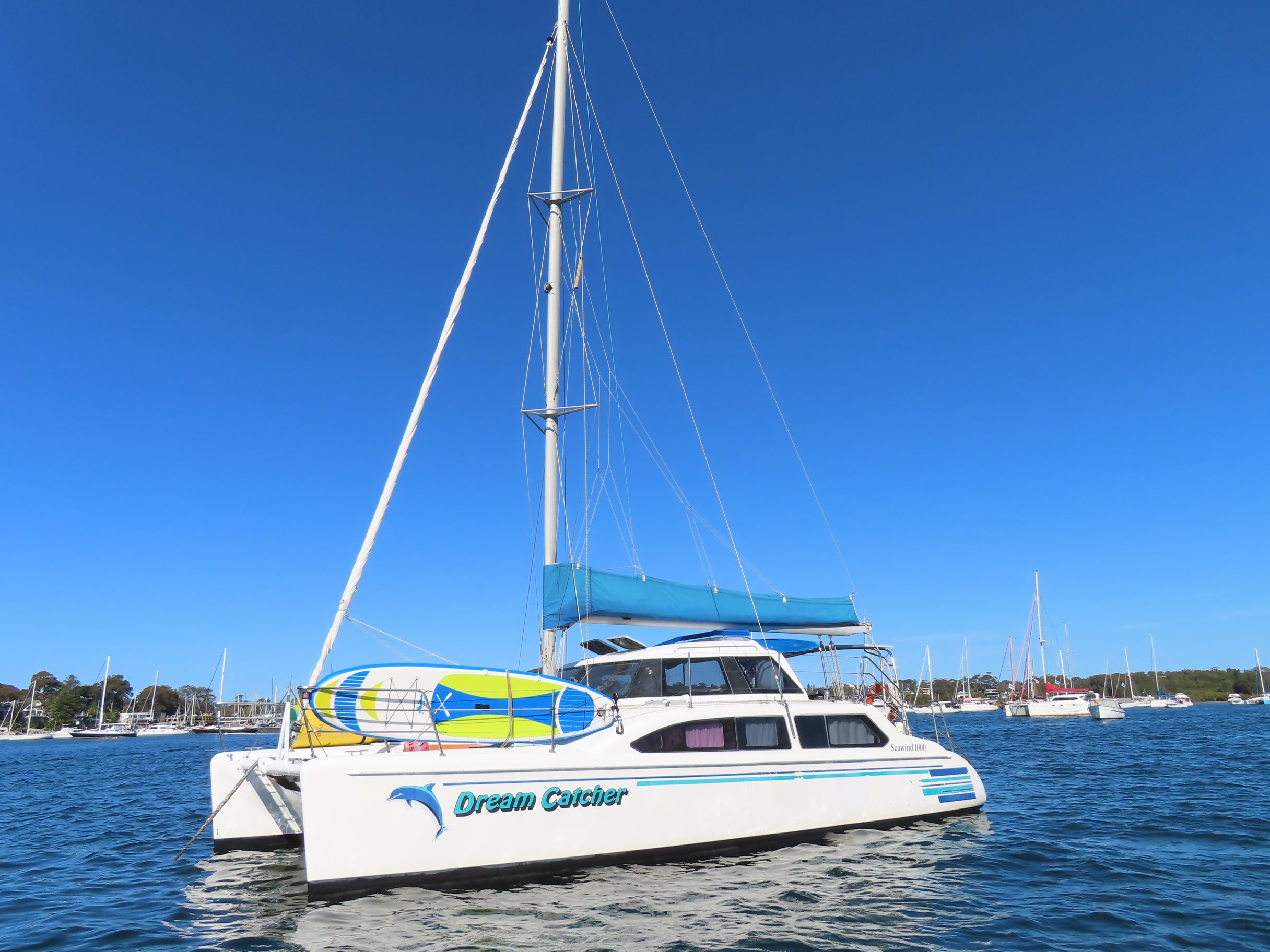 A white catamaran sailboat named "Dream Catcher" floats on calm blue water under a clear sky, with a paddleboard secured on top and other boats visible in the background.