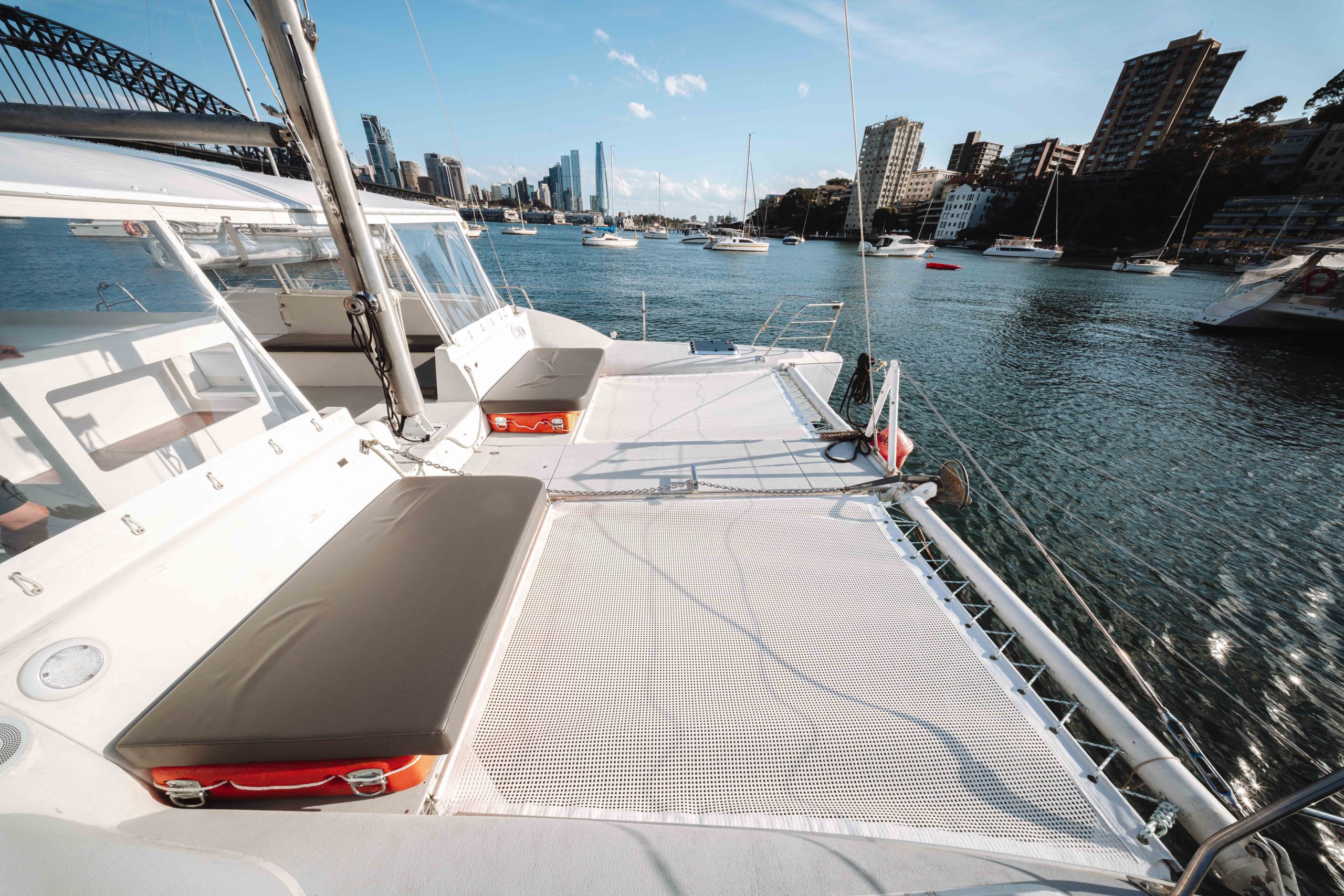 Sunny view from the deck of a catamaran anchored in a harbor, with nearby boats, city buildings, and a bridge in the background under a blue sky with scattered clouds.