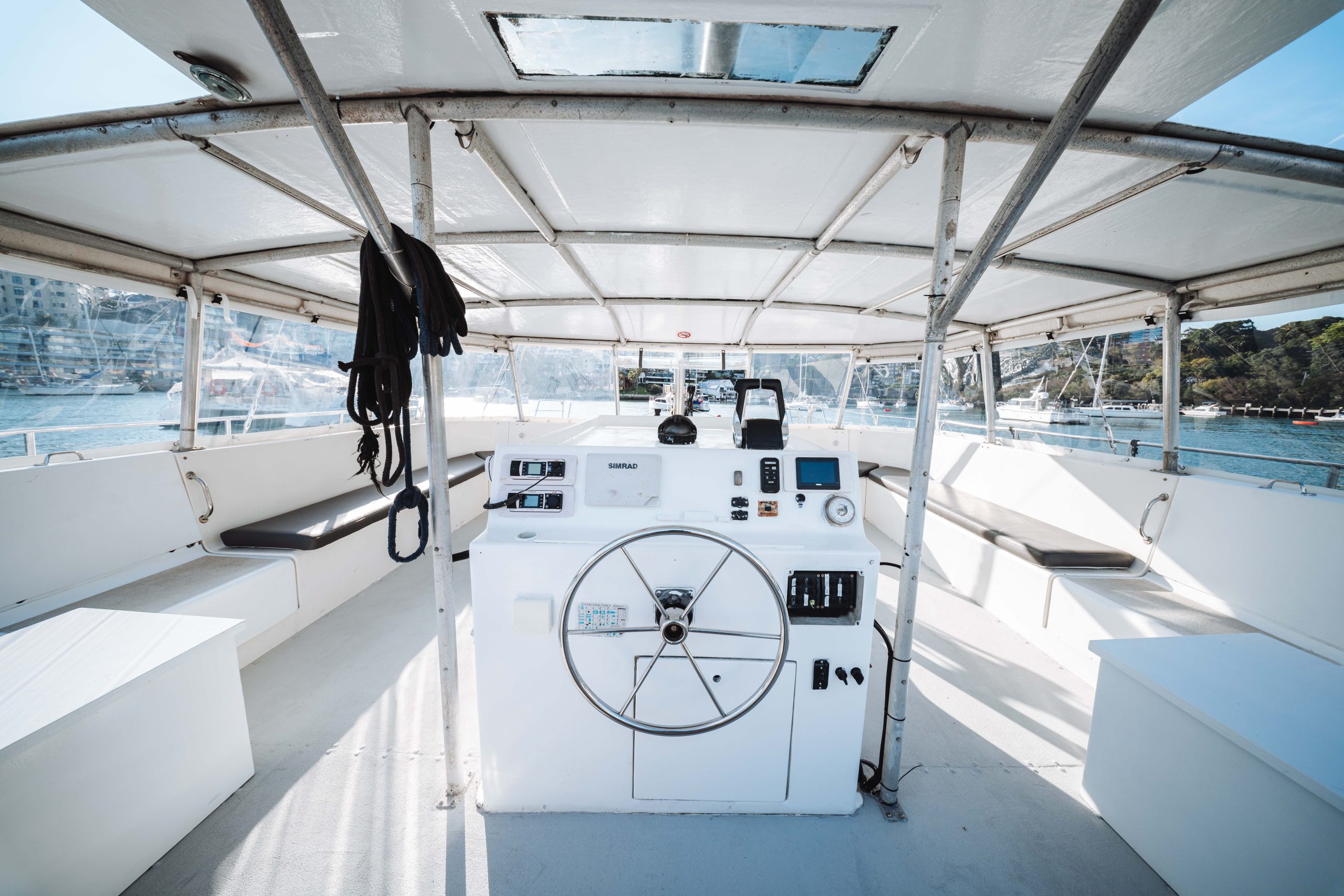 The image shows the steering area of a boat with a large white control panel, a steering wheel, navigational equipment, and seating on both sides. The boat is docked near a marina with water and greenery visible outside.