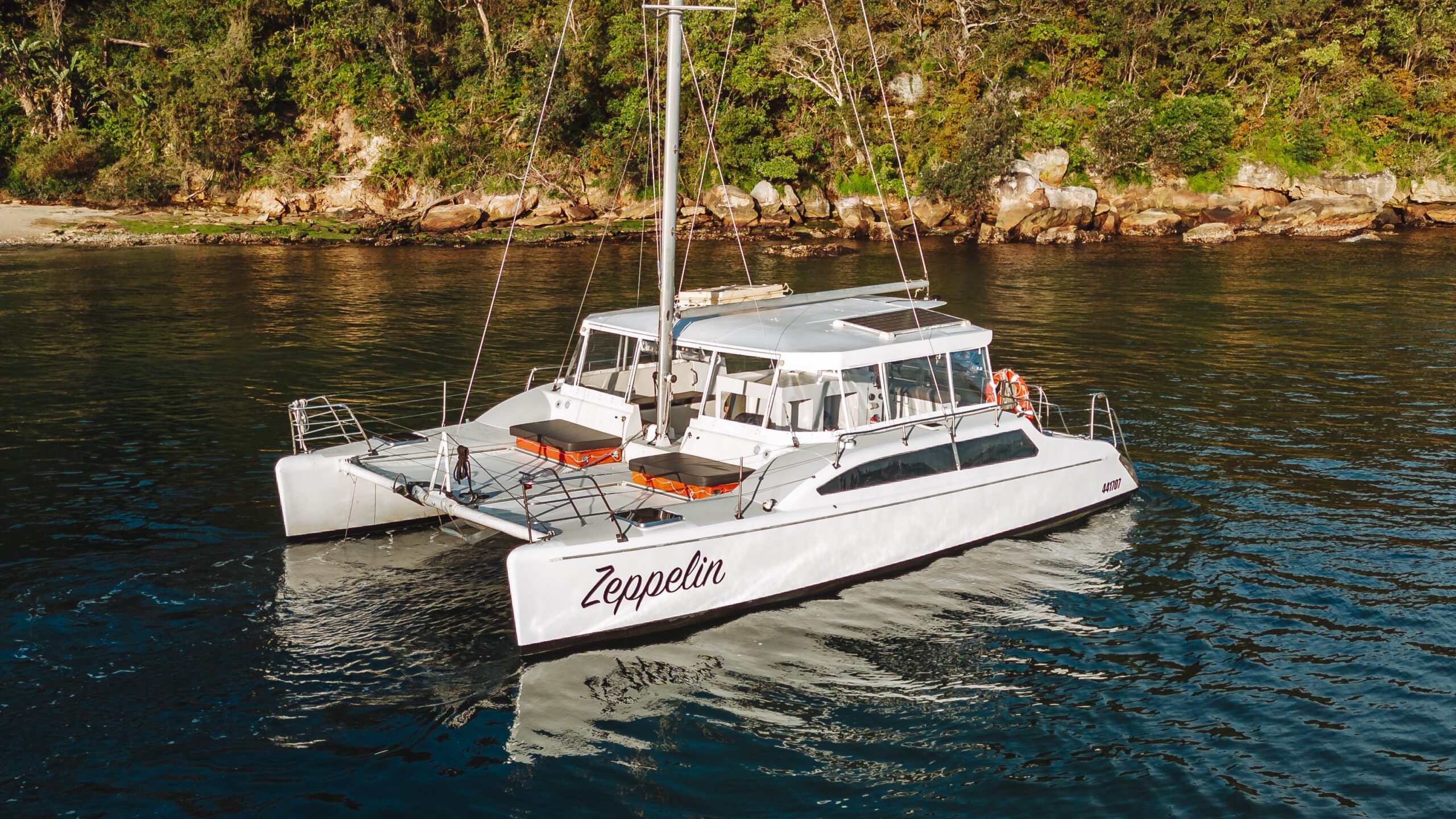 A white catamaran named "Zeppelin" floats on calm water near a rocky shoreline with dense green trees in the background.