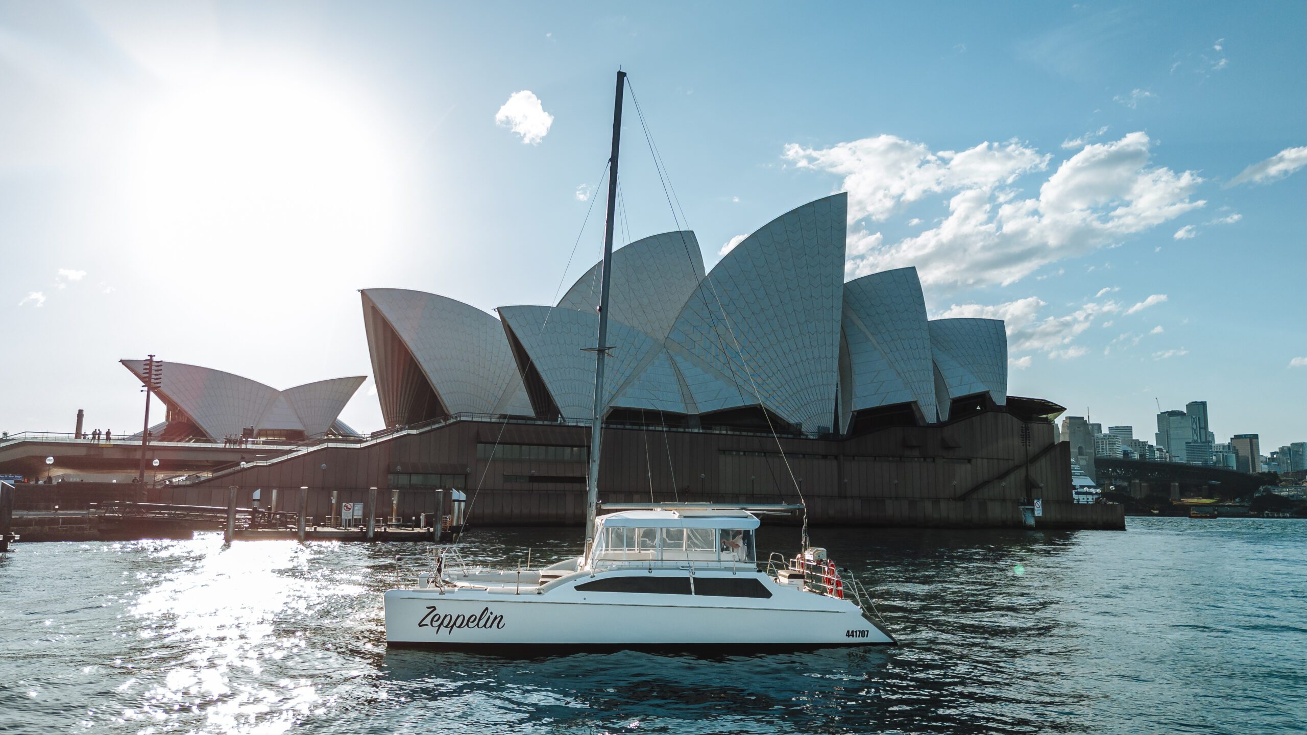 A white sailboat named Zeppelin floats on the water in front of the Sydney Opera House under a bright, sunny sky, with city buildings visible in the background.