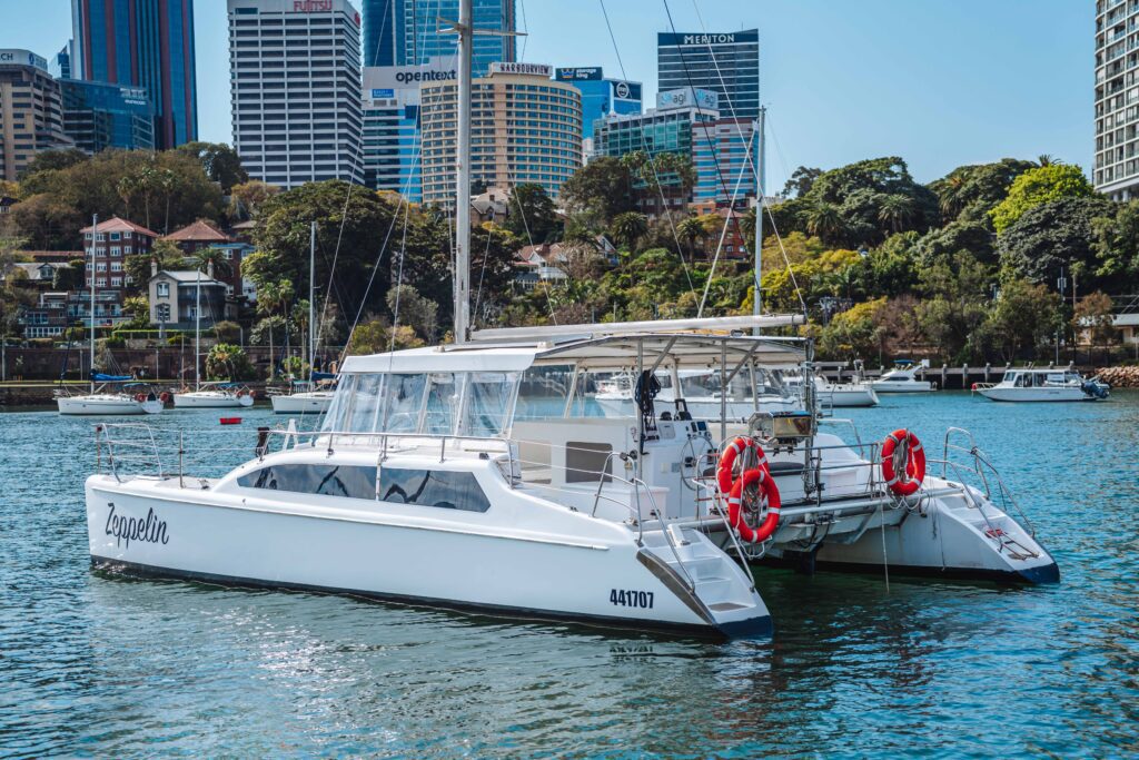 A white catamaran named "Zagalein" floats on a calm river with city buildings and greenery in the background. The boat has red lifebuoys and is moored near other boats under a clear sky.