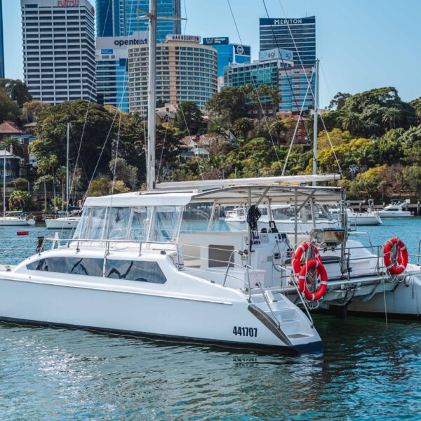 A white catamaran named "Zagalein" floats on a calm river with city buildings and greenery in the background. The boat has red lifebuoys and is moored near other boats under a clear sky.