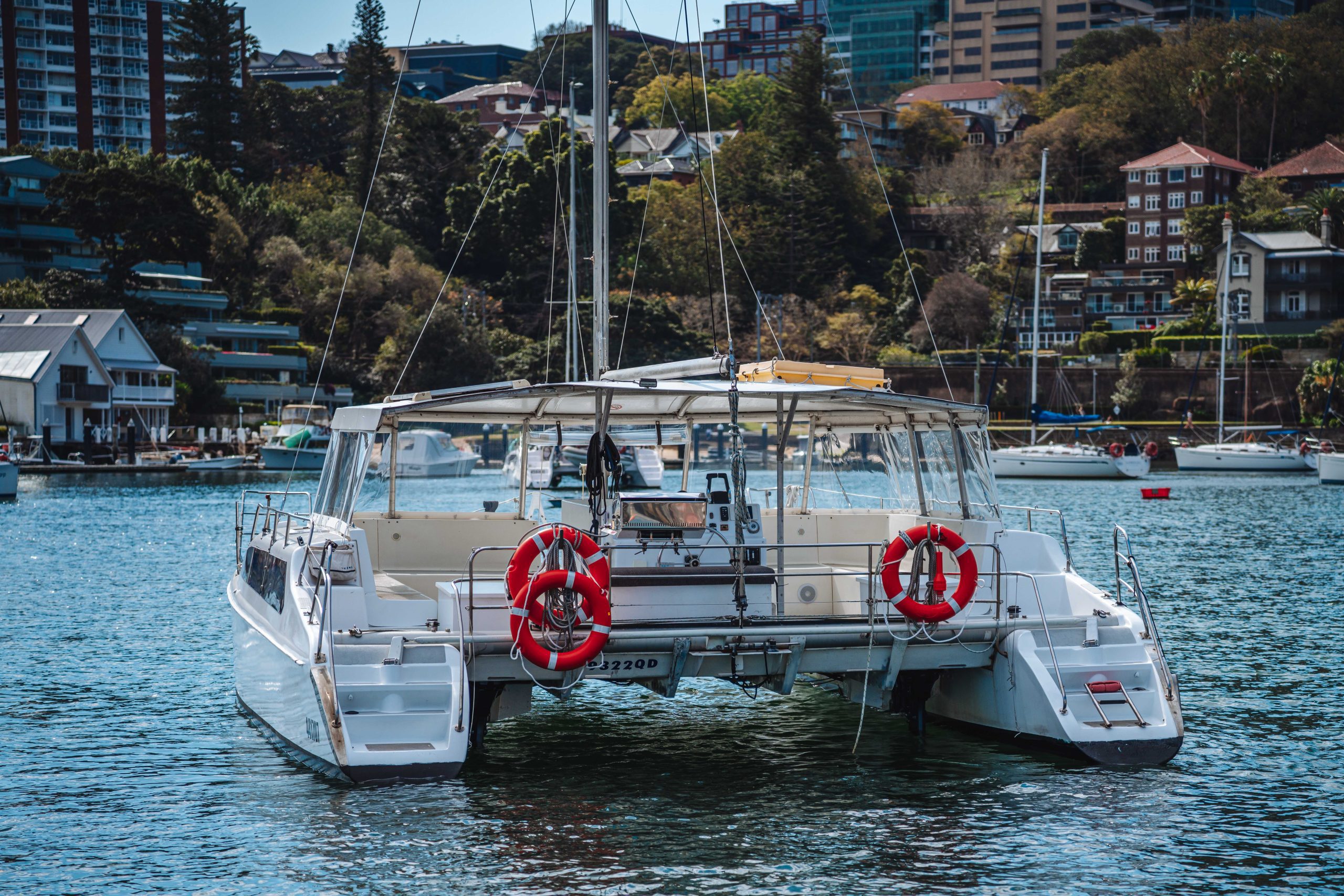 A white catamaran with two red lifebuoys on its back deck is docked in a calm harbor, surrounded by water, other boats, and waterfront buildings with trees in the background.