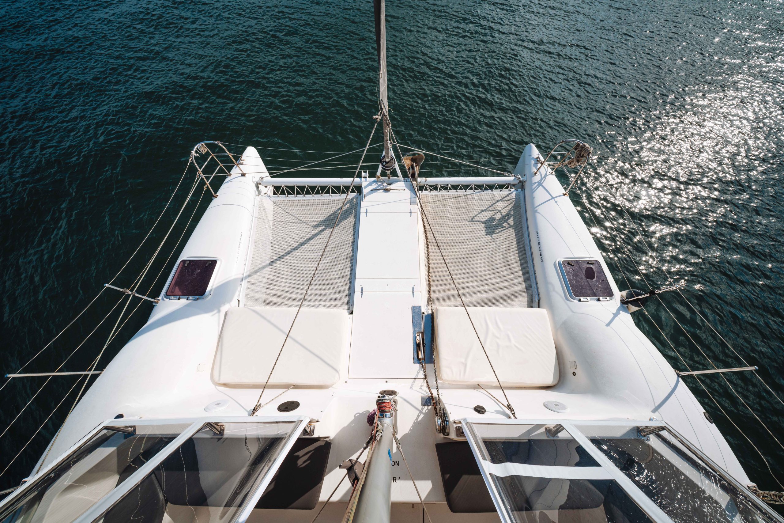 A top-down view of a white catamaran sailboat on calm blue water, showing its twin hulls, netted deck area, and part of the main mast. Sunlight reflects off the rippling water.