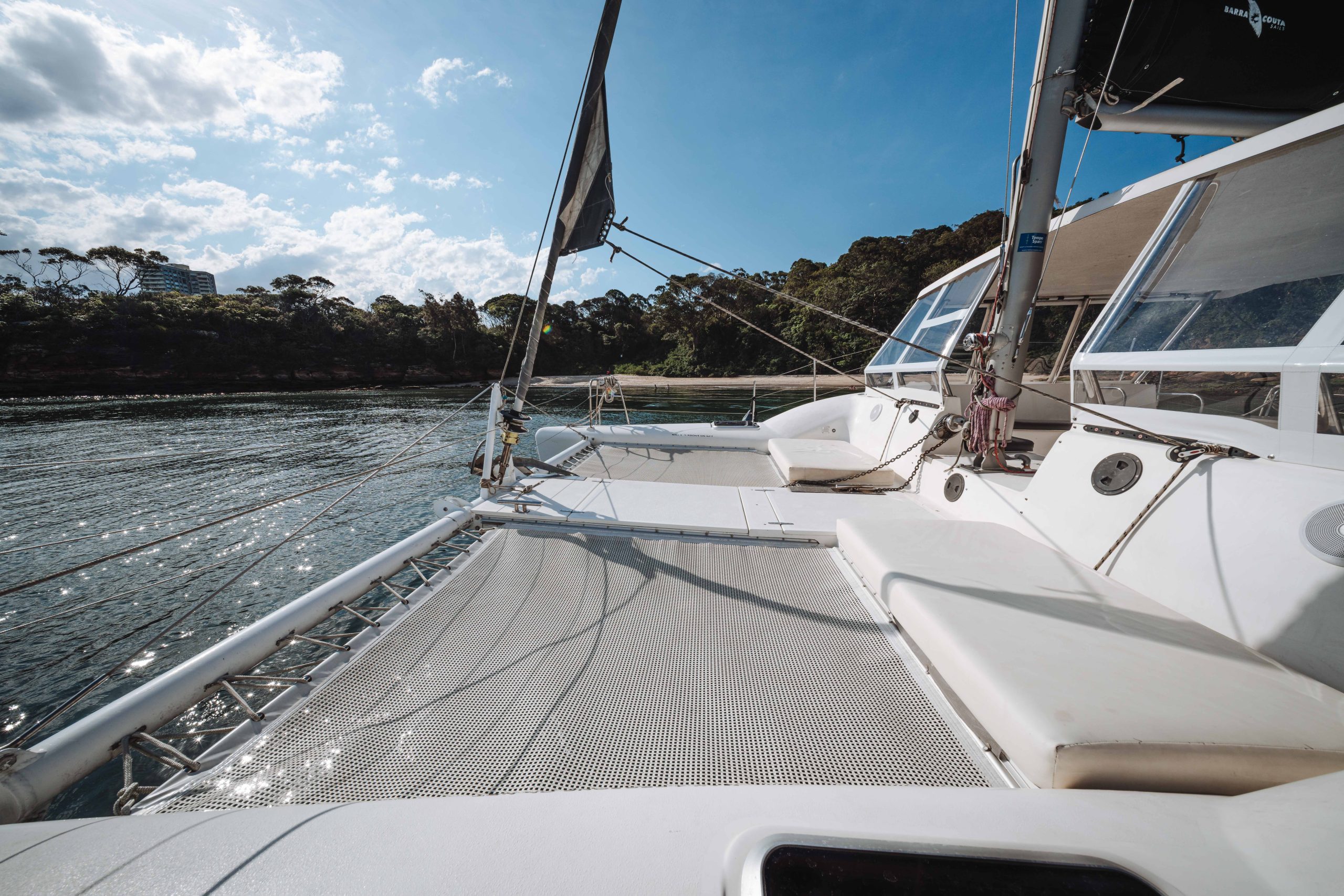 A view from the deck of a catamaran sailboat on calm water, with trees and a partly cloudy sky in the background. The netted area and ropes are visible in the foreground.