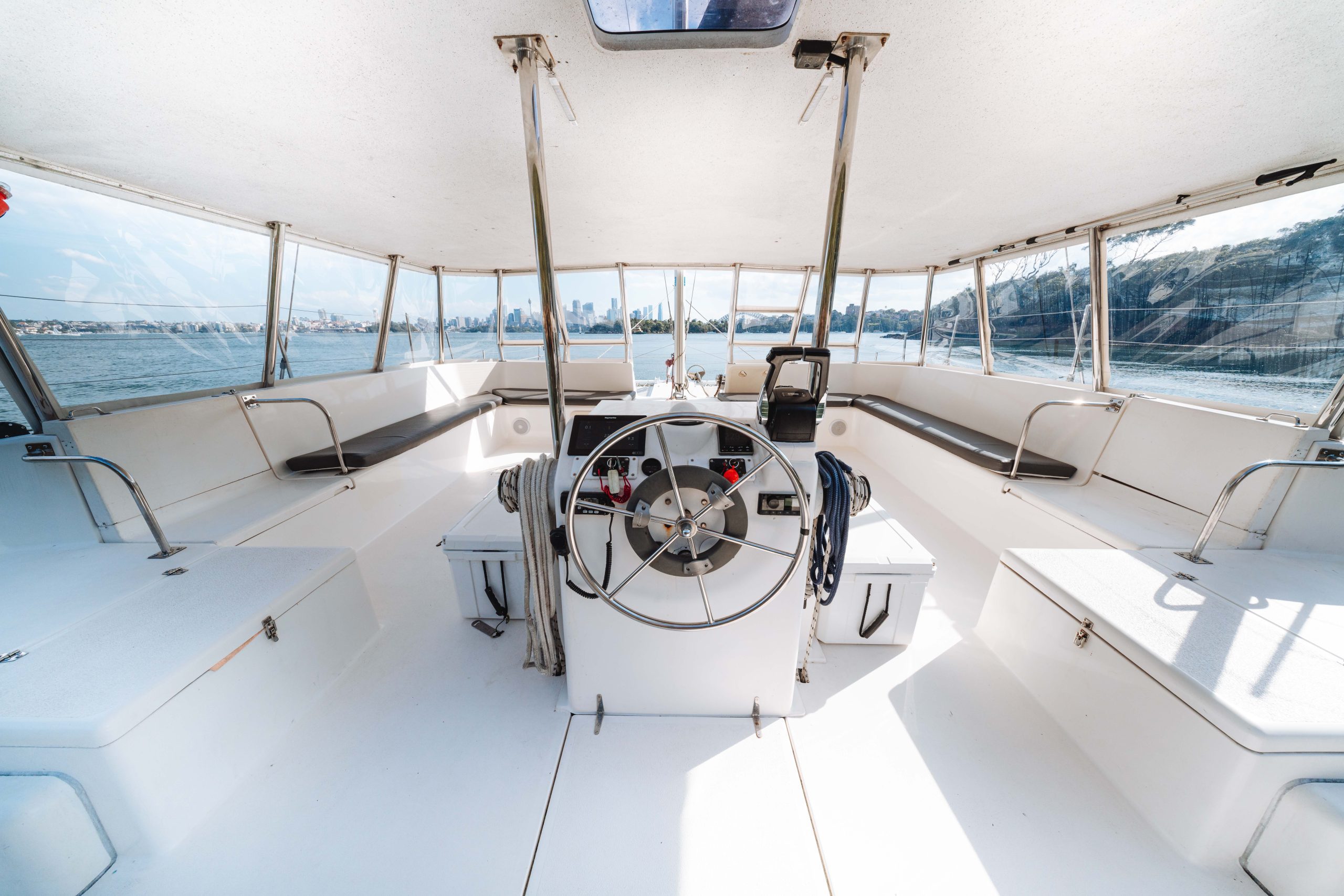 The interior of a boat's cockpit with a central steering wheel, benches along the sides, large windows offering views of the water, and a distant shoreline visible outside.