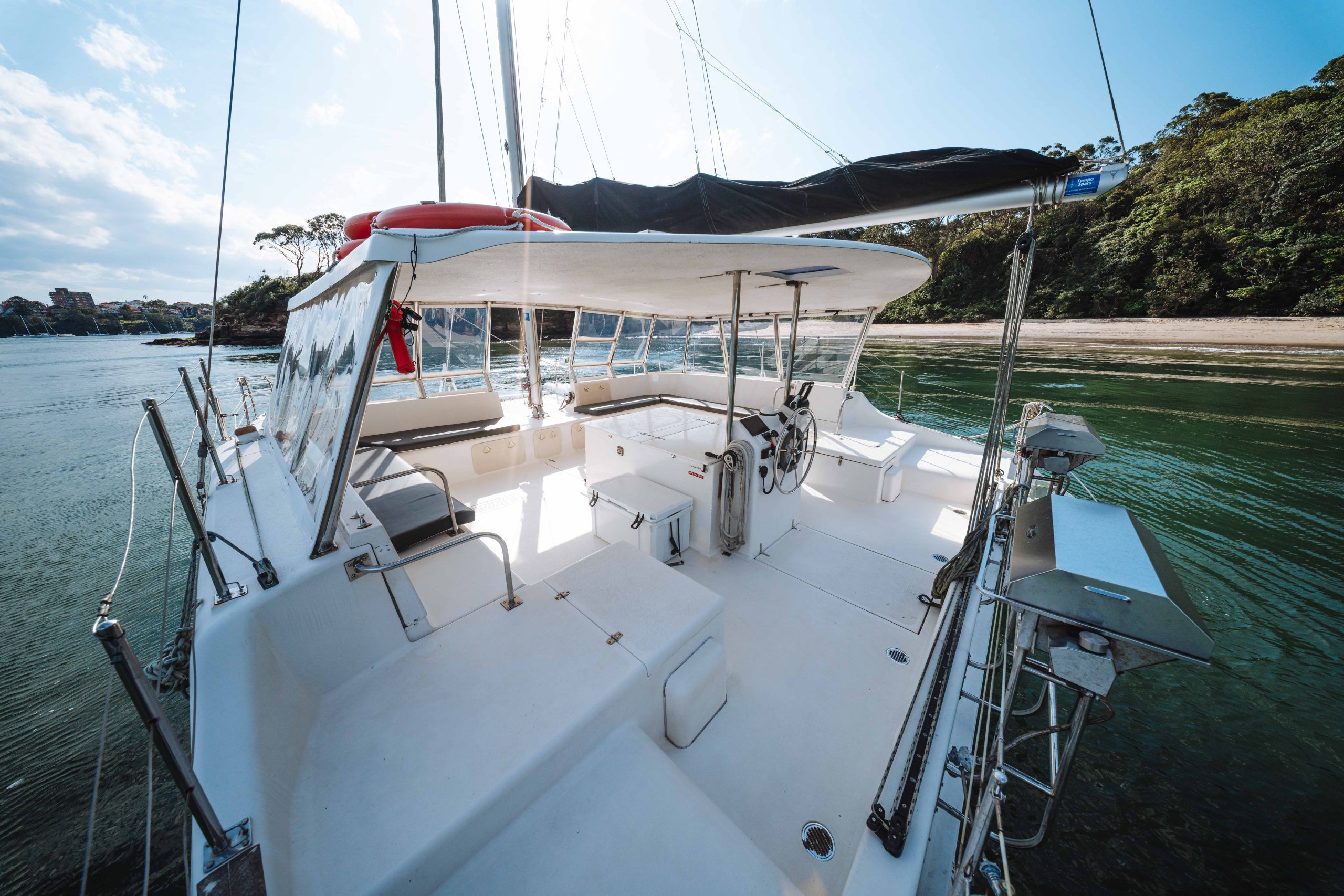 A view from the stern of a white catamaran sailboat anchored near a wooded shore, showing the spacious cockpit with seating, a steering wheel, and surrounding metal railings under a canopy.