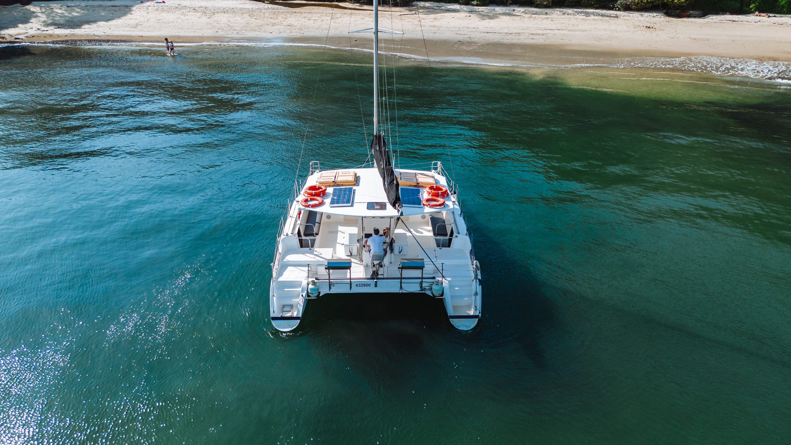 A white catamaran with people on board floats on clear blue-green water near a sandy beach, with gentle waves and a person standing at the shoreline in the background.