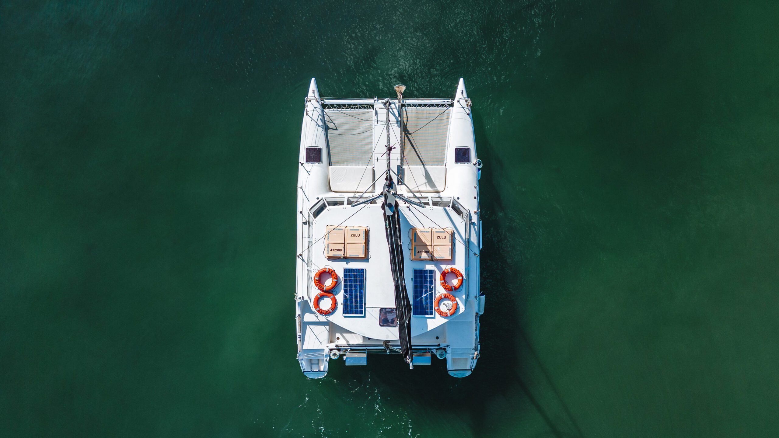 Aerial view of a white catamaran with solar panels and lifebuoys on deck, floating on calm, green water.