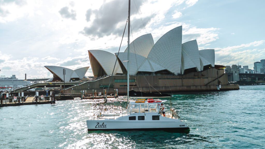A white sailboat named Zulu floats on the water near the Sydney Opera House, with its iconic sail-shaped roof and a cityscape in the background under a partly cloudy sky.