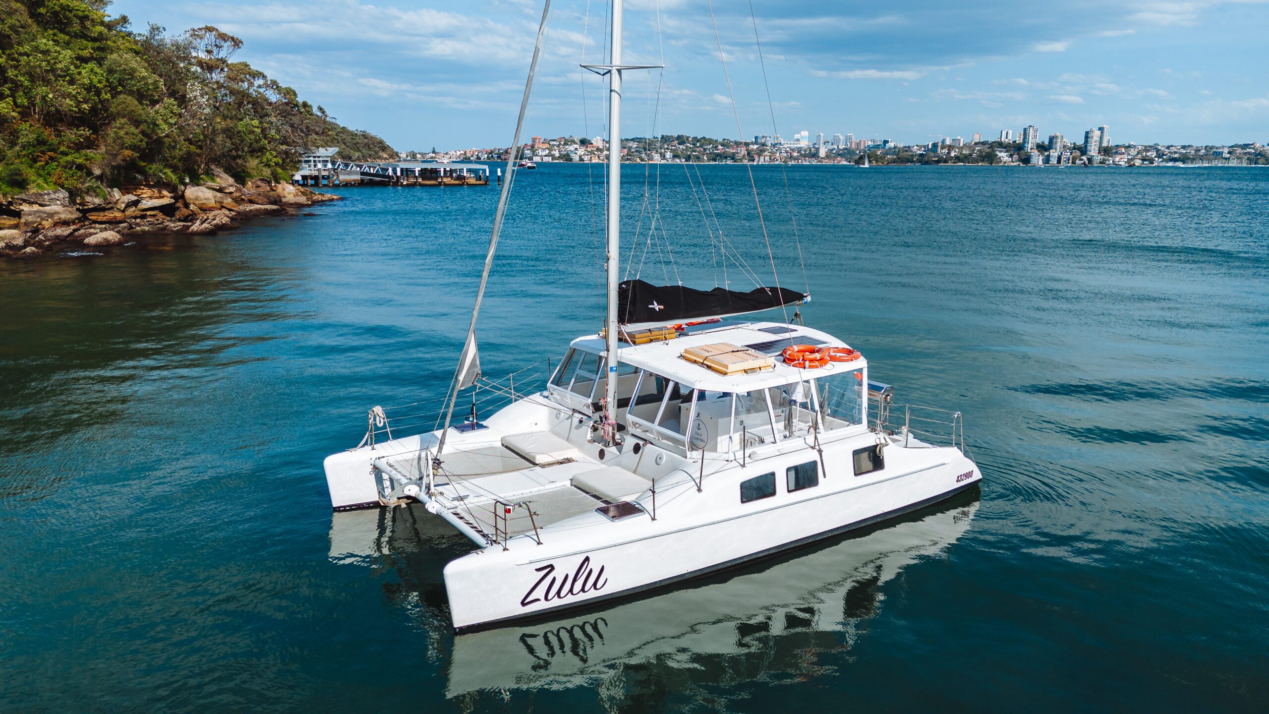 A white catamaran named "Zulu" floats on calm blue water near a rocky shoreline with trees. City buildings and a pier are visible in the background under a partly cloudy sky.