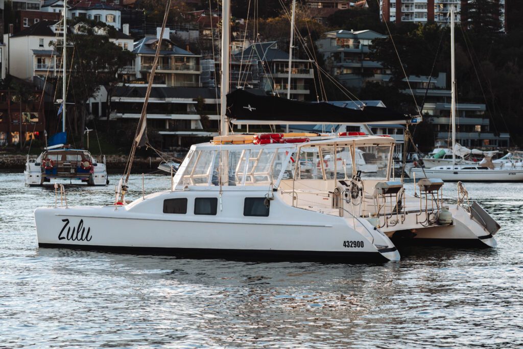 A white catamaran named "Zulu" floats on calm water near other sailboats, with waterfront buildings and trees in the background.