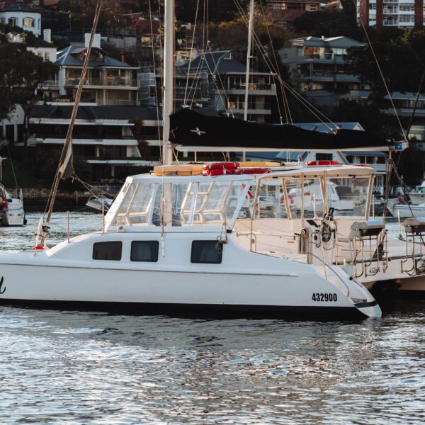 A white catamaran named "Zulu" floats on calm water near other sailboats, with waterfront buildings and trees in the background.