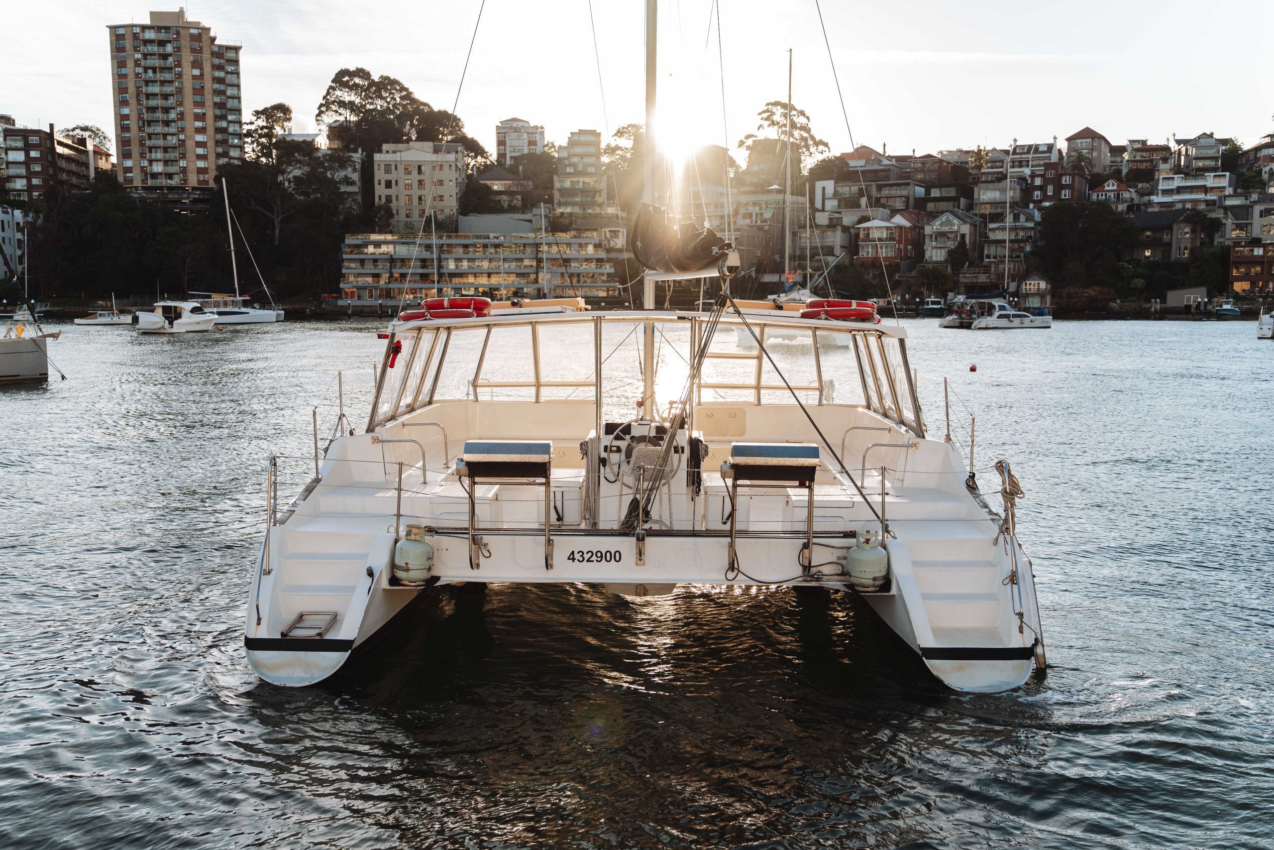 A white catamaran boat docked on calm water, with the sun setting behind it and city buildings and trees visible in the background.