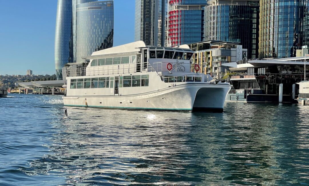 A white multi-deck yacht floats on calm water near a modern city waterfront with tall glass skyscrapers and clear blue sky in the background.
