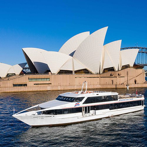 A white tour boat sails on the water in front of the iconic Sydney Opera House, with its distinctive white sail-like roof structures and clear blue sky in the background.