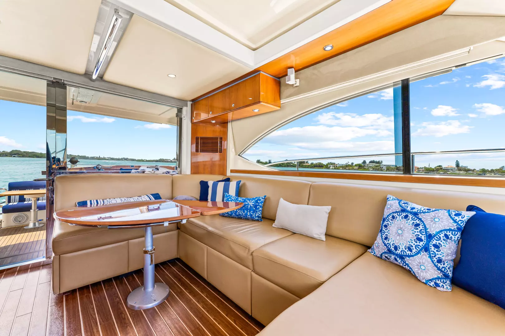 Luxurious yacht interior with beige leather seating, blue and white throw pillows, a wooden table, large windows, and views of water and shoreline under a partly cloudy sky.