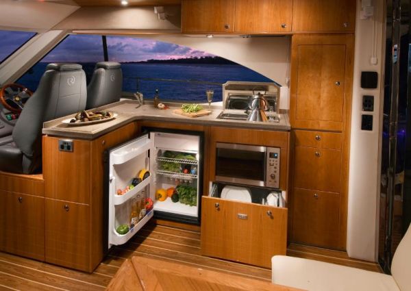 A compact boat kitchen with wooden cabinets, a sink, a small stove, an open fridge stocked with food, and a dishwasher drawer. Large windows show a scenic sunset over water in the background.