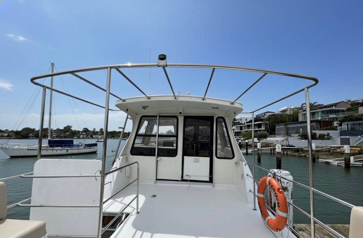 A view from the deck of a docked white motorboat, showing the cabin, metal railings, and a life ring, with waterfront houses and other boats in the background under a clear blue sky.