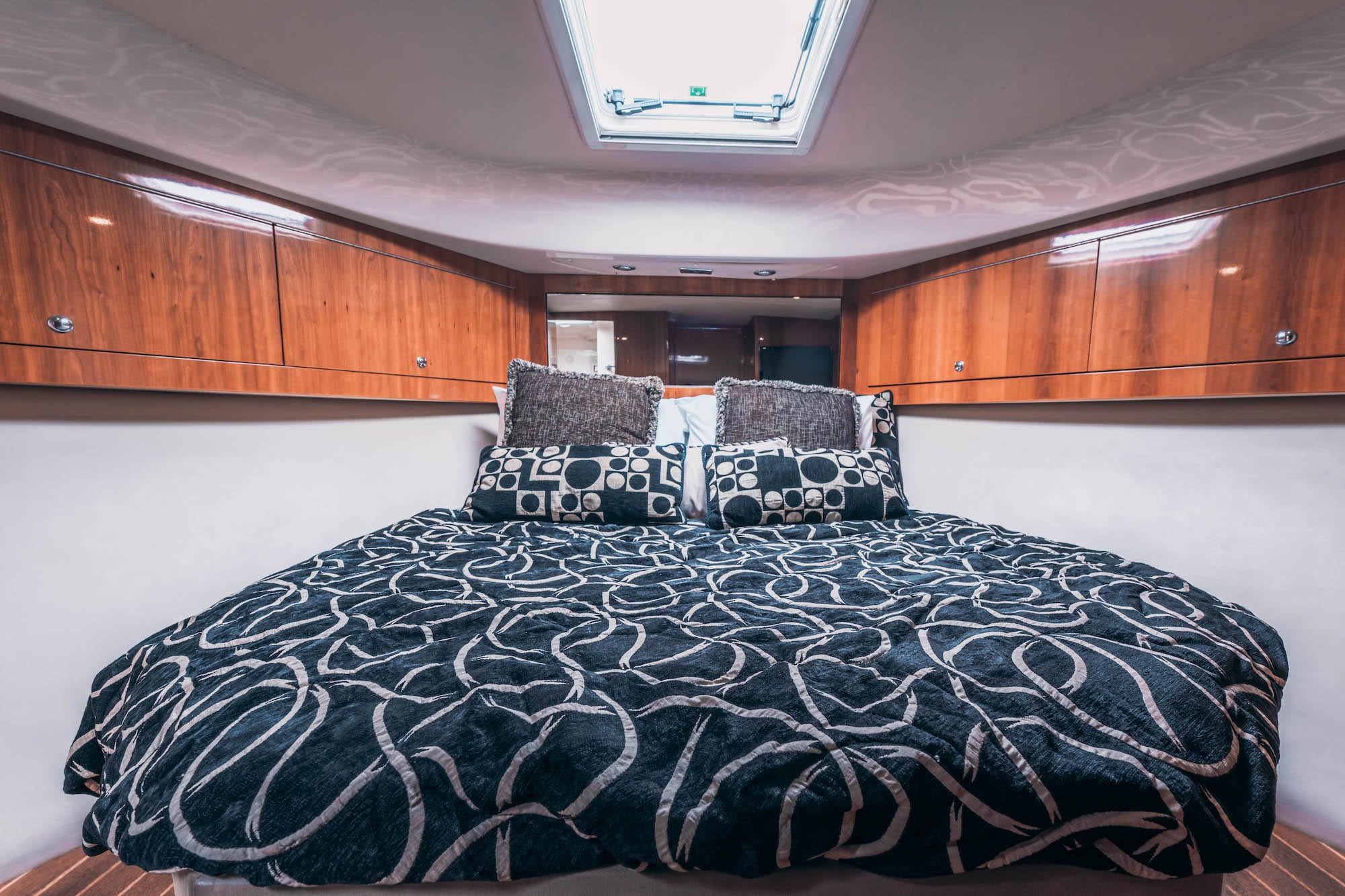A cozy boat cabin bedroom with a round bed covered in a black and white abstract-patterned comforter, matching pillows, wooden cabinets above, and a ceiling hatch letting in natural light.