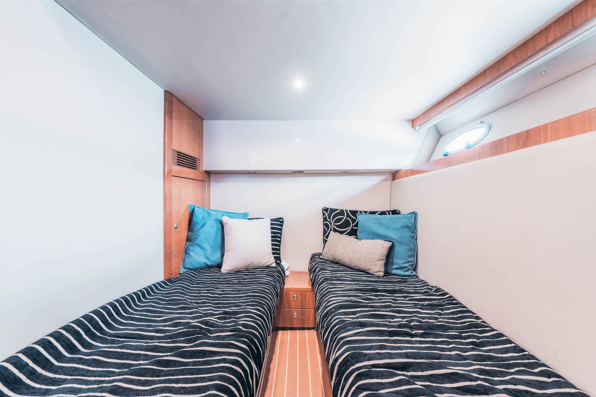 A cozy cabin room with two single beds, each with striped bedding, colorful cushions, and a small wooden nightstand in between. Light wood accents and a small window create a bright, inviting space.