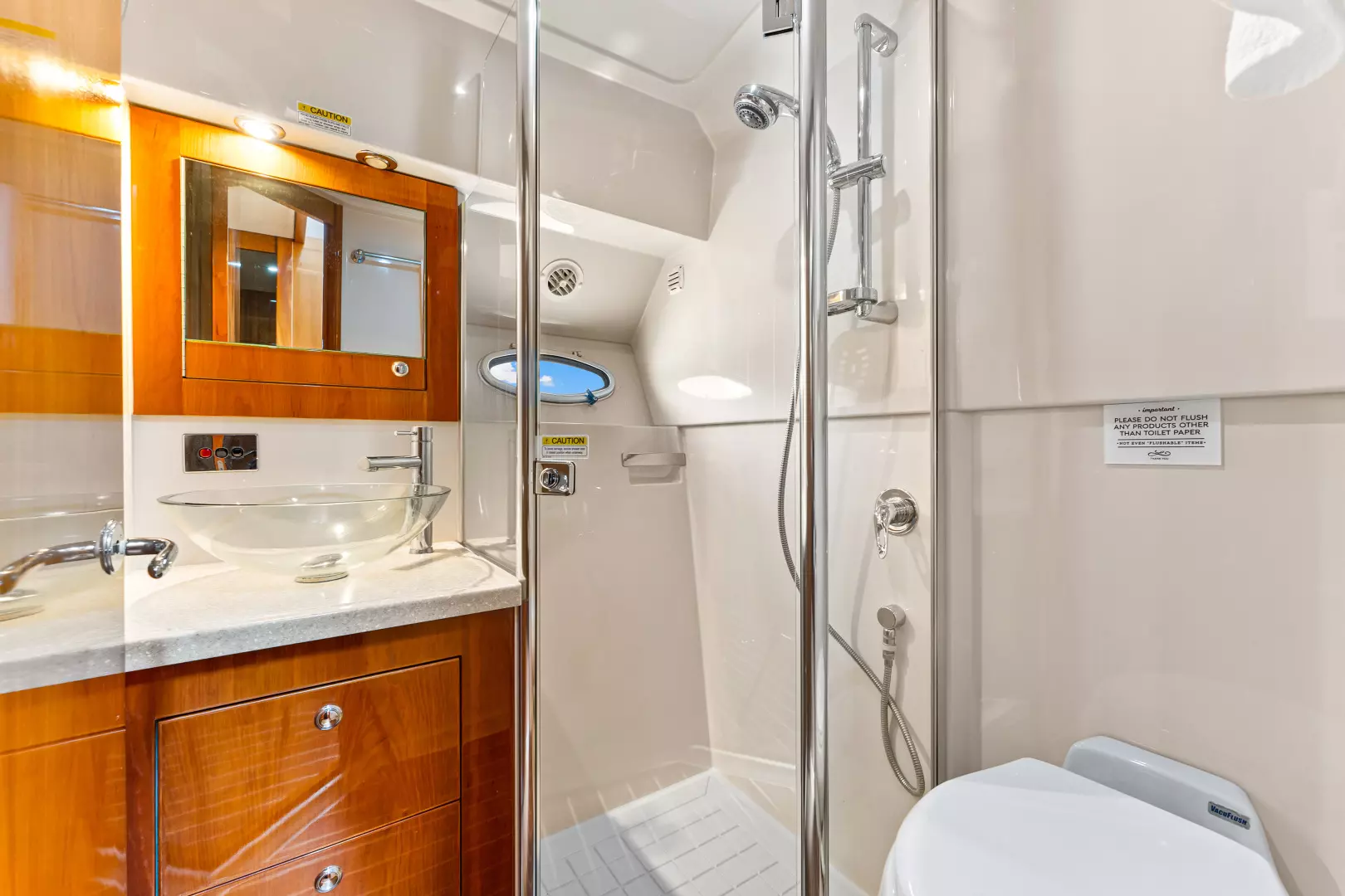 Modern bathroom with wooden cabinets, a glass vessel sink, a wall-mounted mirror, a shower with glass doors, chrome fixtures, and a white toilet. Small round window above the sink. Bright and clean space.