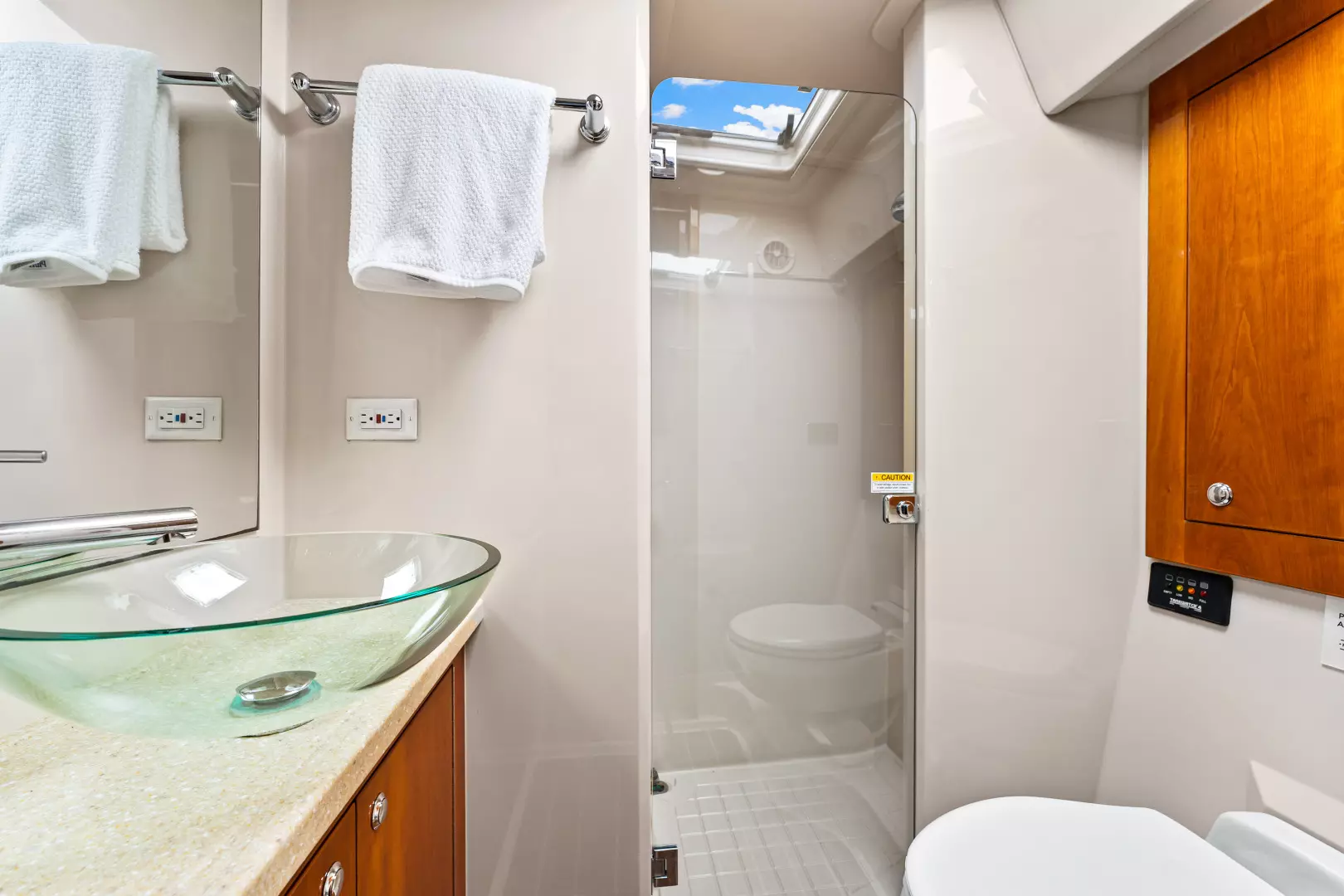 Modern bathroom with a glass vessel sink, wooden cabinets, a glass-enclosed shower, white towels on racks, and a skylight letting in natural light.