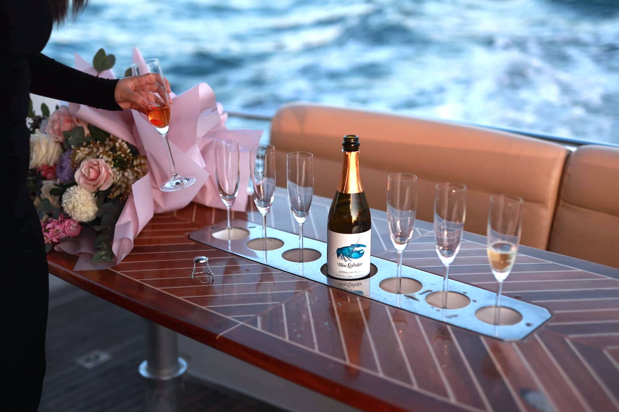 A person holds a champagne glass next to a bouquet of flowers, six empty glasses, and a champagne bottle on a wooden table aboard a boat, with water visible in the background.