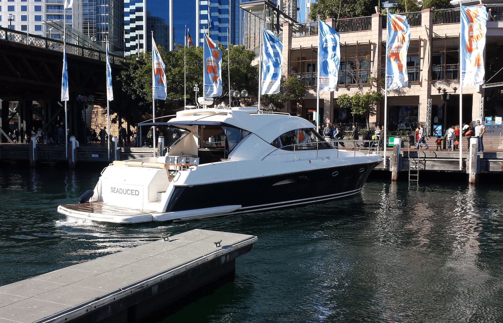 A luxury yacht named "Sea-Duced" is docked in a city harbor, with modern buildings, colorful flags, and people walking along the waterfront in the background.