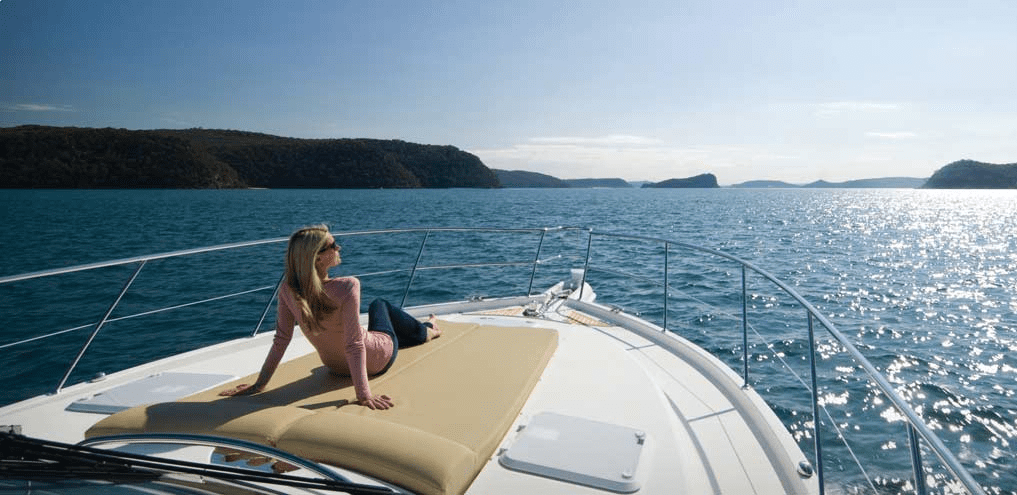 A woman relaxes on a yacht's sun pad, looking out over calm blue water with forested hills and islands in the background under a clear, sunny sky.