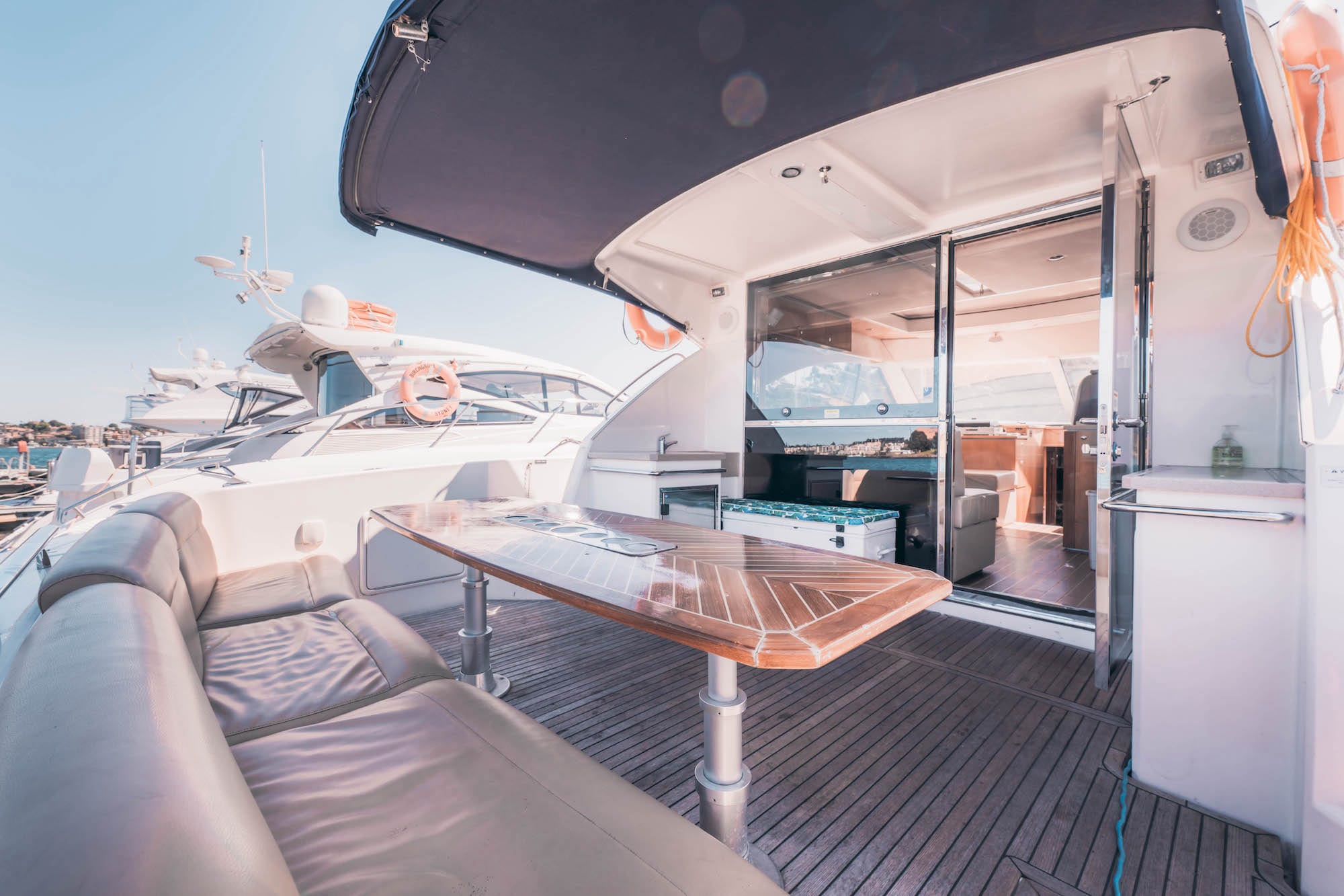 A luxurious yacht deck with a cushioned seating area and a wooden table under a canopy, with sliding glass doors leading to an interior lounge and other yachts visible in the marina background.