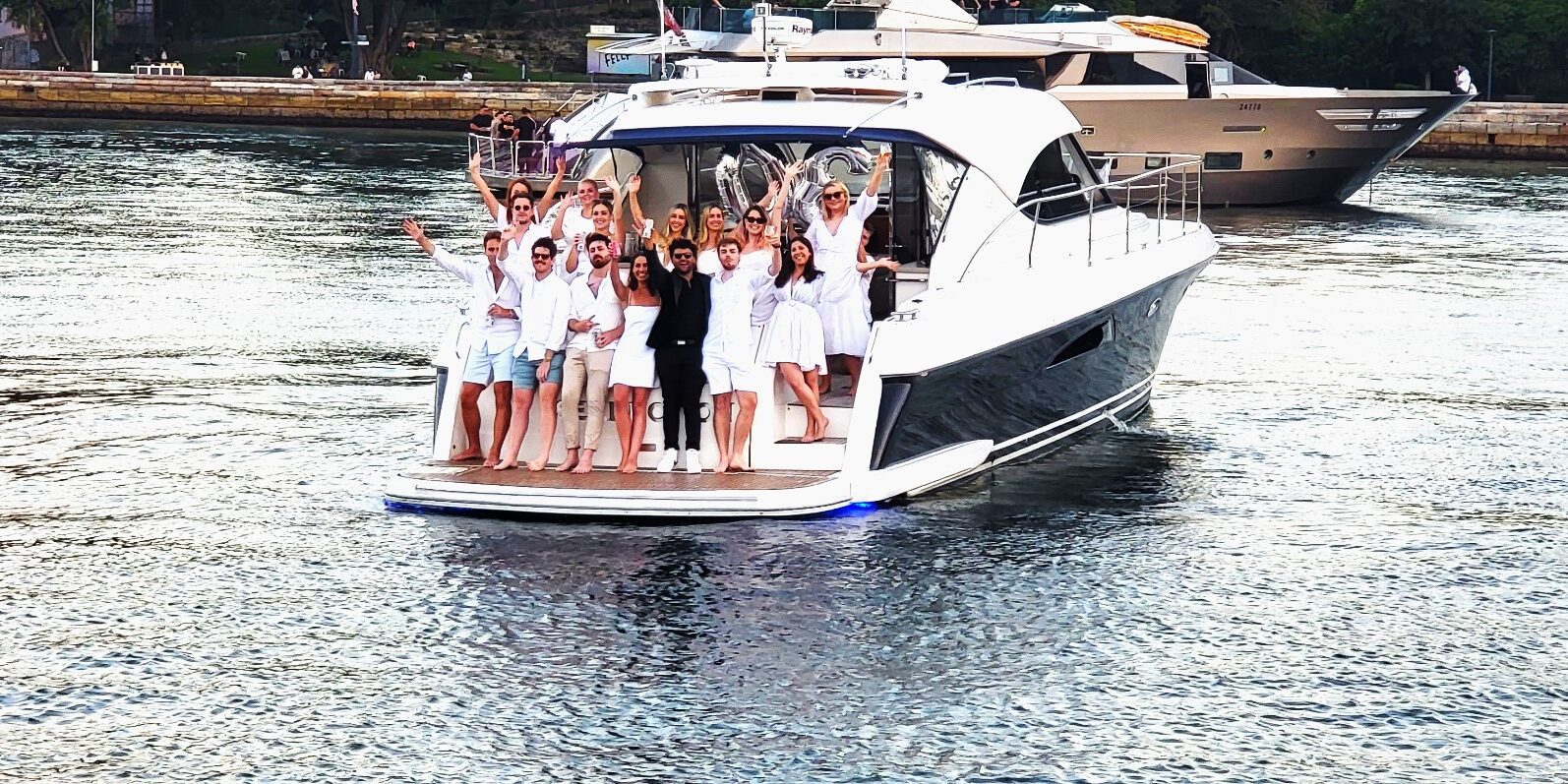 A group of people dressed in white and black outfits stand and wave from the back deck of a yacht on the water, with another yacht and a stone dock visible in the background.