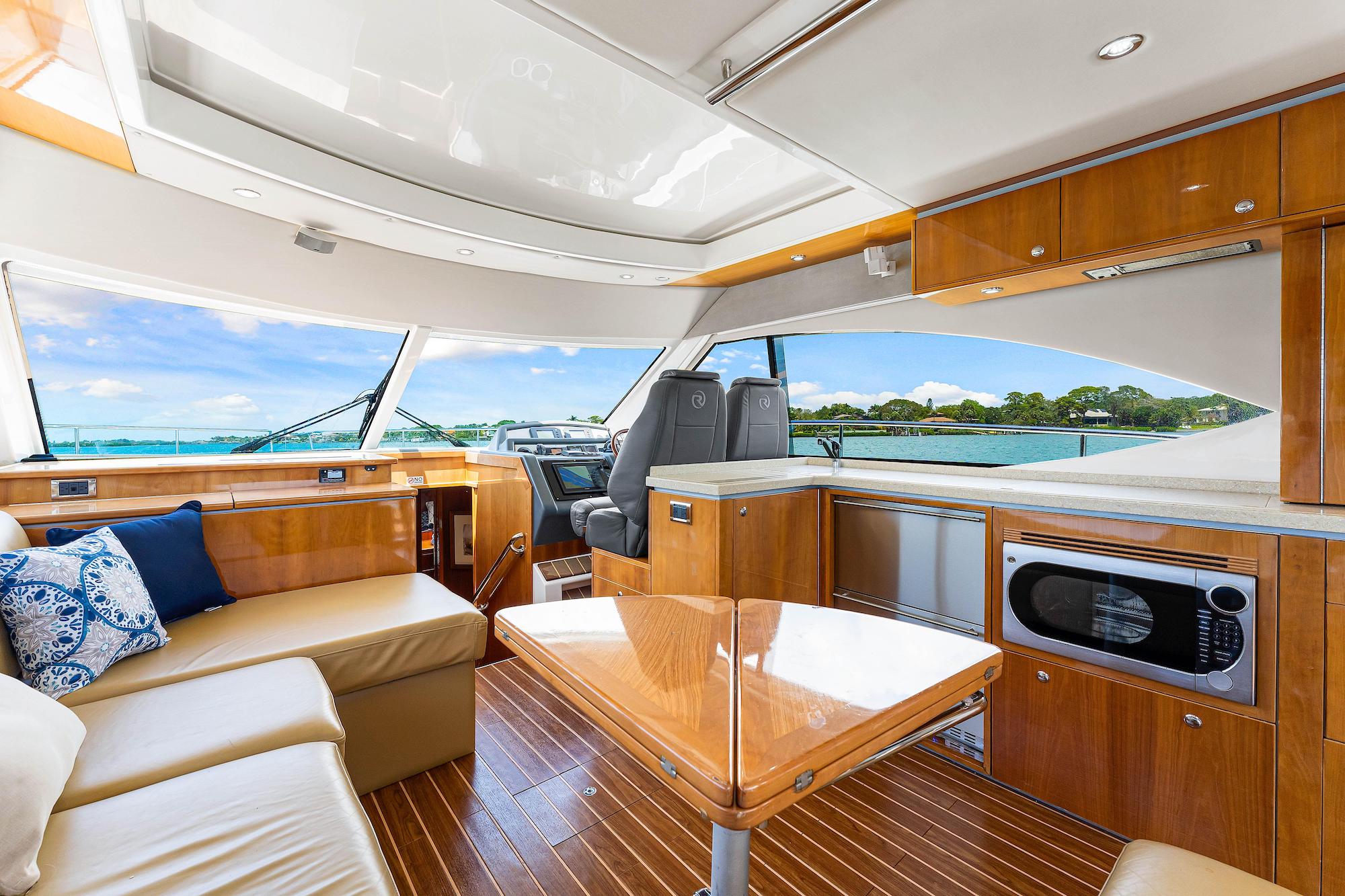 The interior of a modern yacht features tan seating with blue patterned pillows, a glossy wooden table, a kitchenette with a microwave, and large windows offering a scenic water view.