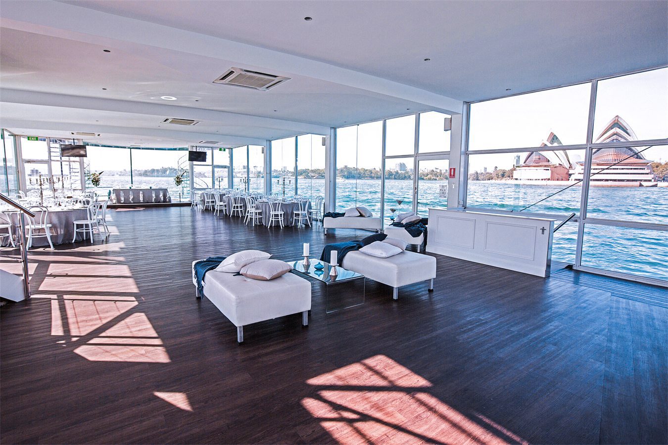 Spacious, modern event venue with large windows overlooking water and the Sydney Opera House, featuring white furniture, dark wood floors, and abundant natural light.